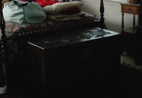 A nicely carved wooden chest on a Persian carpet sits at the foot of a four post bed with a nice wooden end table in the background. There is an assortment of blankets on the bed.