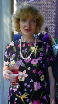 Nora stands with a drink in a dark dress with big color splotches.