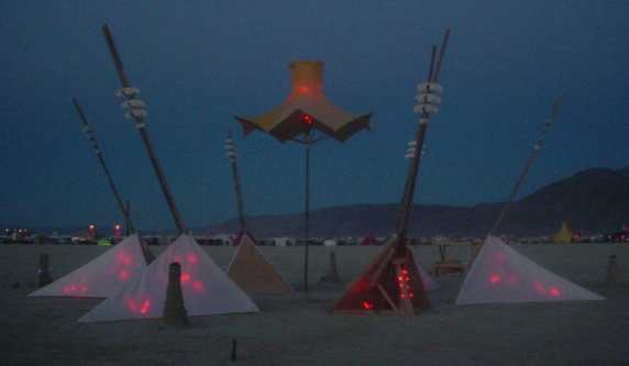 As dawn begins to lighten the sky, a light sculpture with shapes stands out against the playa with mountains in the background.