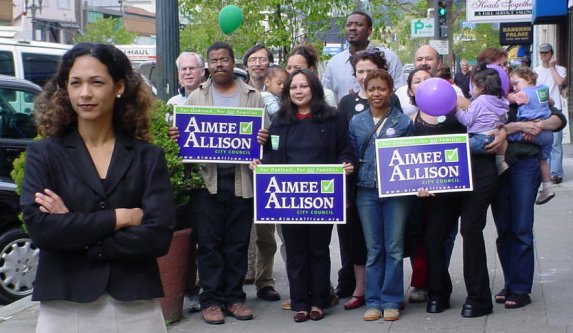 Aimee Allison stands in front of a bush, to the left of her supporters who wave "Aimee Allison for City Council" signs. The picture was taken on Grand Avenue in Oakland.