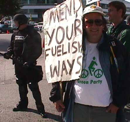 Tian in a Green Party T shirt holds a MYFW sign and stands in front of a policeman in riot gear.