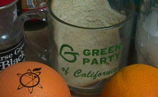 A Green Party of California coffee cup with white bread
          in it sits to the right of some pepper, between two oranges,
          one real and one fake.