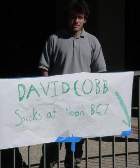 Justy with a sign that says "DAVID COBB speaks here at noon" with an arrow pointing toward room B67.