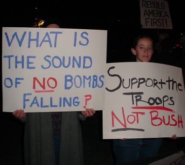 Two people holding signs. One says "What is the sound of no bombs dropping?".