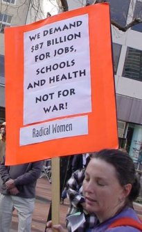 Woman carrying a sign that says "we demand that 87 billion be spent on us here."