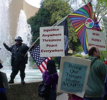 Batman mugs by a water fountain while other people with signs parade past.