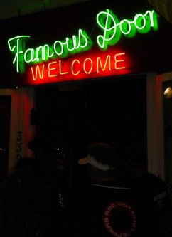A Black guy watches in front of a doorway with "Famous Door" in green neon light above it.