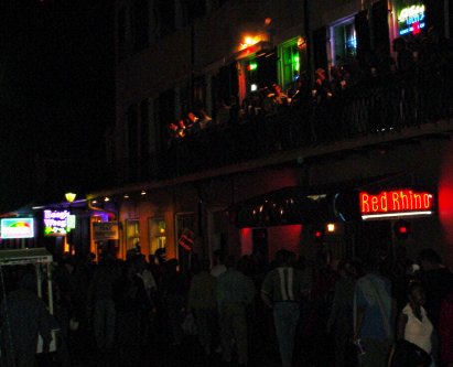 People mill around on Burbon Street. Above them, more people mill around on a balcony. In the background the neon lights are bright.