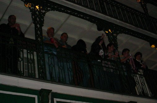A woman gestures as if she has just thrown a strand of Mardi Gras beads off a balcony where she stands with many others.
